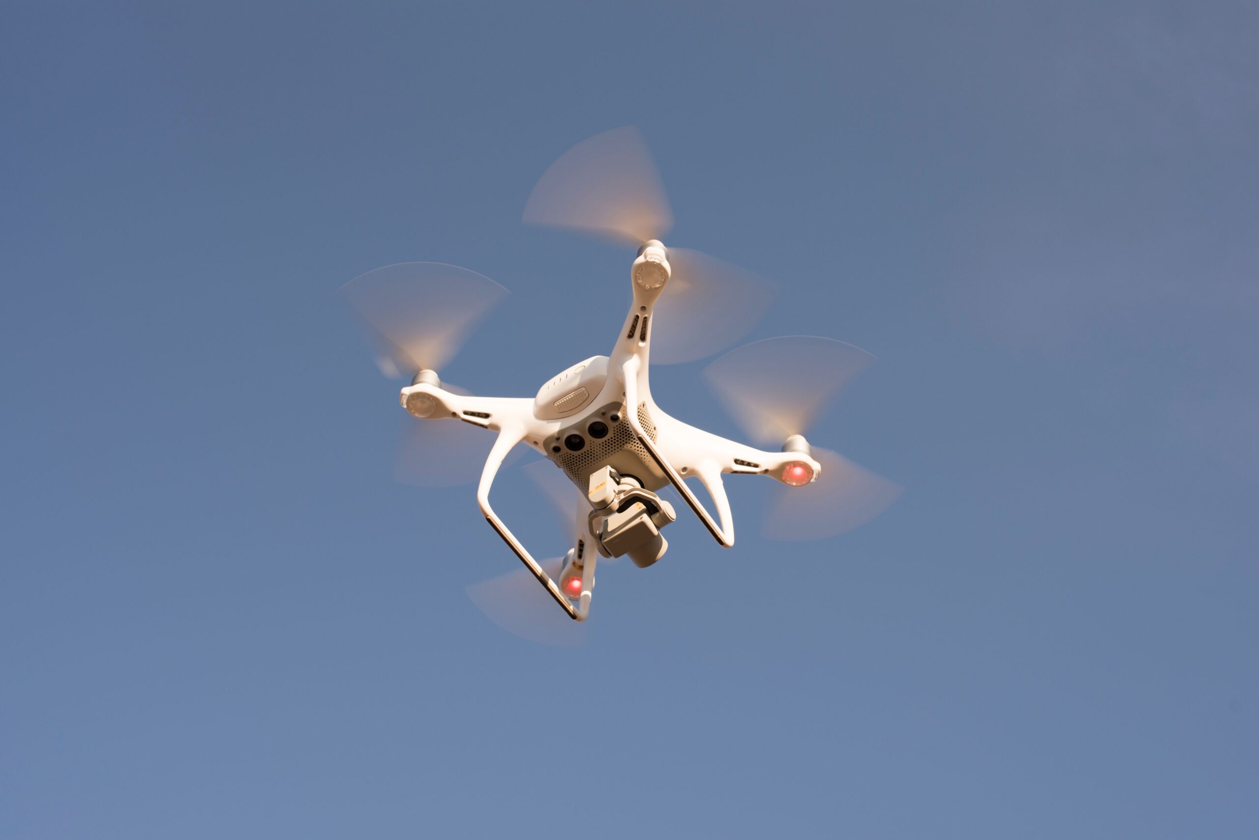 Drone quadricóptero branco em voo, visto de baixo, com hélices em rotação e luzes vermelhas acesas contra um céu azul limpo.
