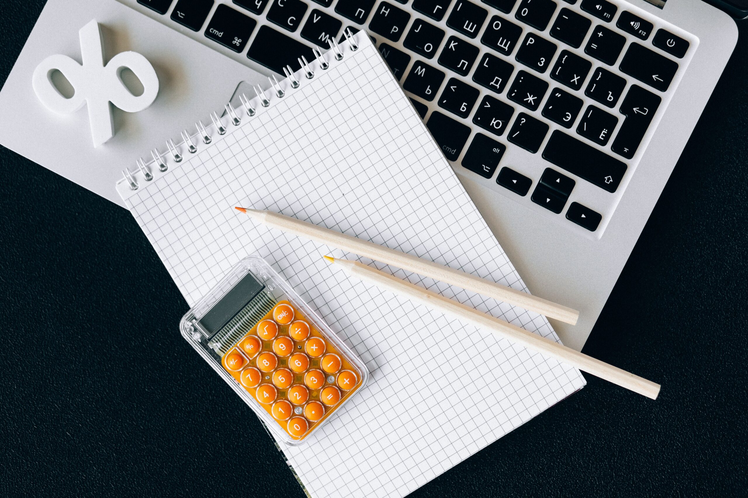 Top-down view of a silver laptop, grid spiral notepad, two wooden pencils, a small calculator with orange buttons, and a 3D percentage symbol, all on a black surface.