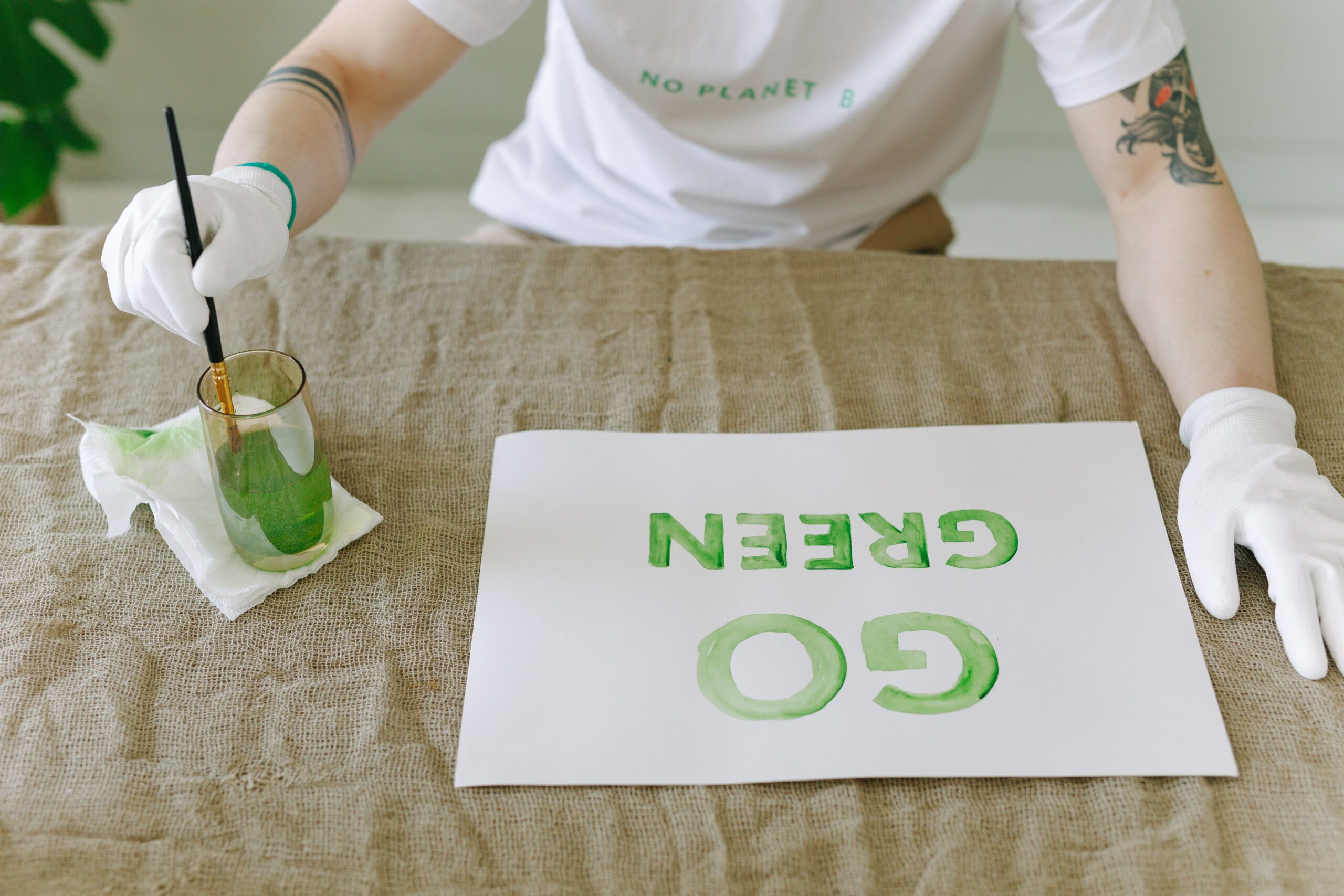 Pessoa usando luvas brancas e camiseta com a frase "No Planet B" segura um pincel ao lado de um copo com tinta verde e um papel onde está escrito "GO GREEN".
