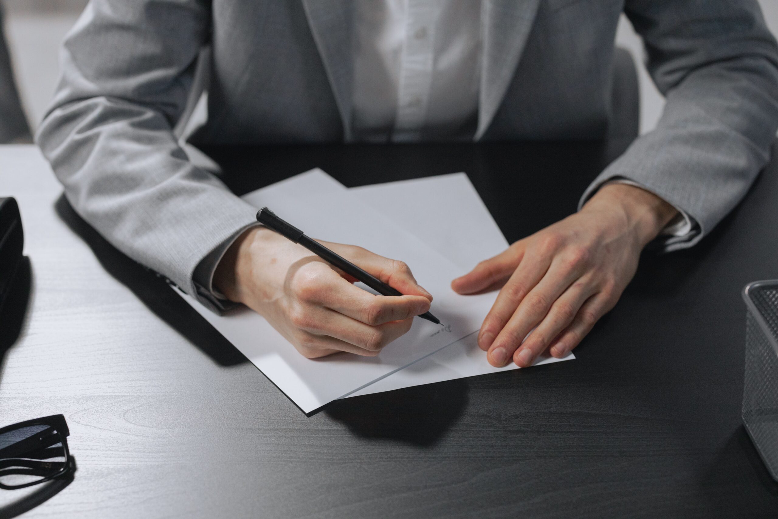 Plano aproximado das mãos de um profissional vestindo terno cinza enquanto redige ou assina documentos sobre uma mesa escura.
